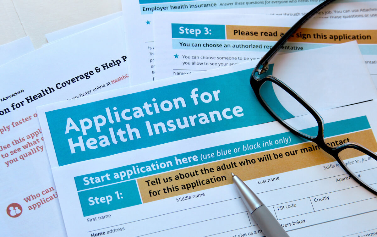 Paper applications for health insurance coverage, arrayed on a surface with a pen